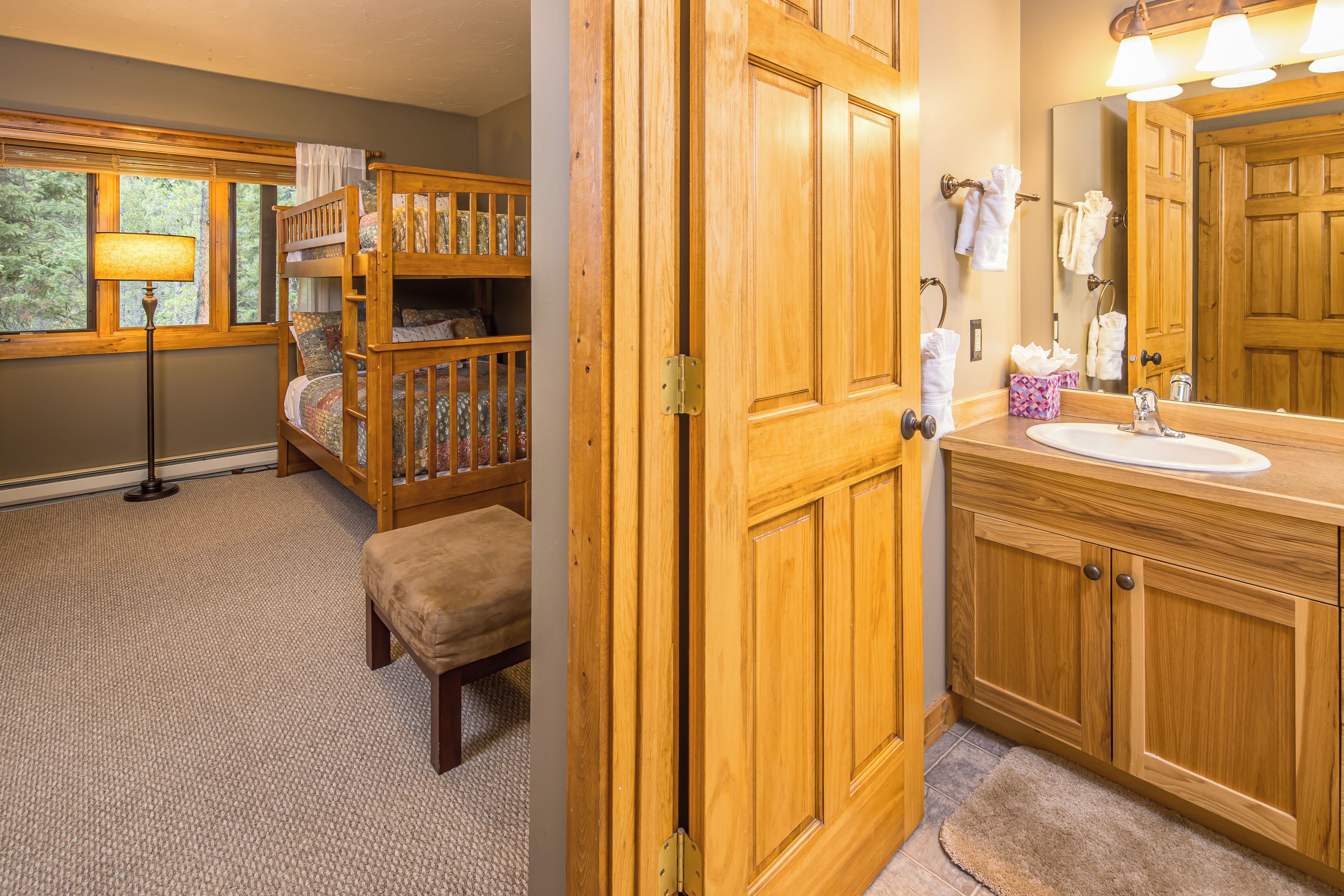 A room with a bunk bed, a bedside lamp, a wooden door, a bathroom with a sink, mirror, and towel rack.