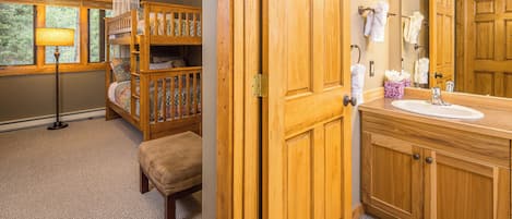 A room with a bunk bed, a bedside lamp, a wooden door, a bathroom with a sink, mirror, and towel rack.