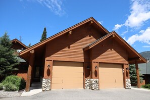 A two-story wooden cabin with a garage, stone accents, and a clear blue sky. - Big Sky Condo! Private Hot Tub & Sauna! Mountain Views! (Big Sky)