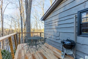 Eine Holzterrasse mit einem Metalltisch und -stühlen, einem Grill und Blick auf Bäume.