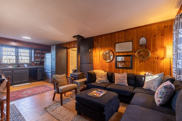 Un salon chaleureux avec un canapé aux couleurs sombres, un mur décoratif en bois et un coin cuisine visible à travers une porte ouverte.