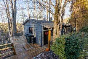 Une terrasse en bois avec une petite table et des chaises, un chalet en bois avec une porte, et un grand arbre en arrière-plan.