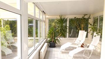 Ein sonniger Balkon mit weißen Stühlen, Topfpflanzen, großen Fenstern und einem gefliesten Boden.