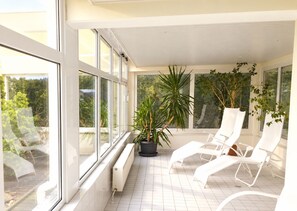 A sunlit balcony with white chairs and potted plants, large windows, and a tiled floor.