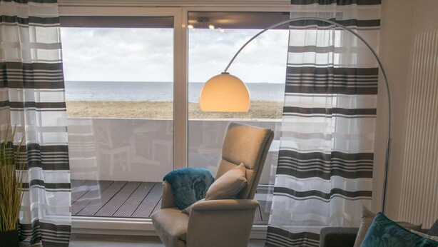 Ein Zimmer mit Blick auf den Strand, ein Sessel, eine Stehlampe und gestreifte Vorhänge.