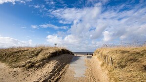 Ein sandiger Pfad, gesäumt von grasbewachsenen Dünen, der zu einem Strand unter einem teils bewölkten Himmel führt.