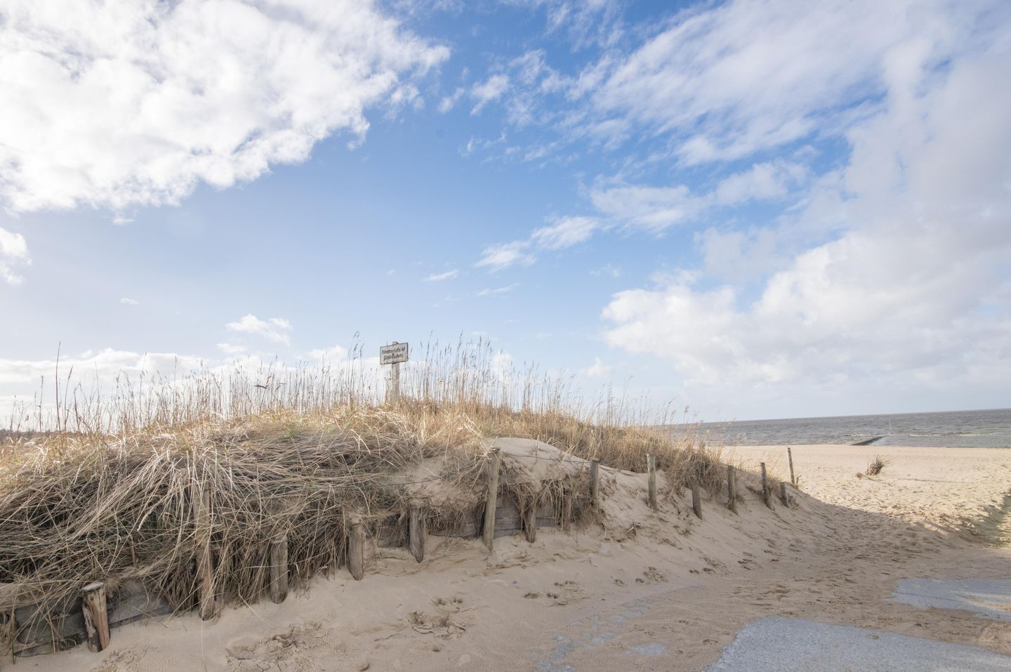 Ein Sandstrand mit Dünen und einem Wegweiser in der Ferne.