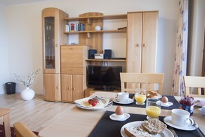 Un coin repas avec une table mise pour le petit-déjeuner, comprenant des assiettes avec des œufs, des toasts et des fruits. On y trouve également un téléviseur et un meuble contenant des livres et des objets décoratifs.