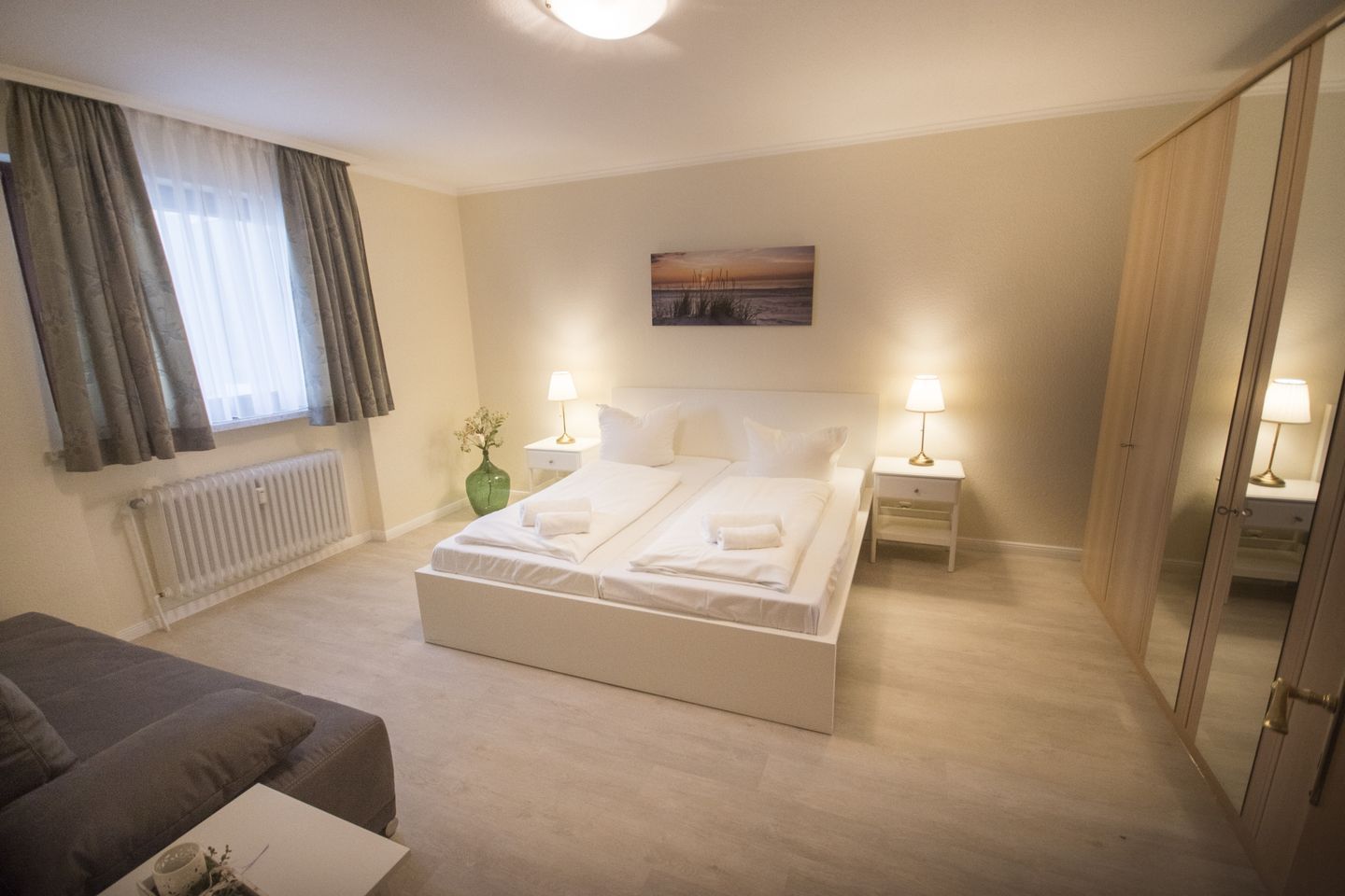 A hotel room with a large bed, two bedside tables with lamps, a radiator, a green vase, a painting on the wall, and a wardrobe.