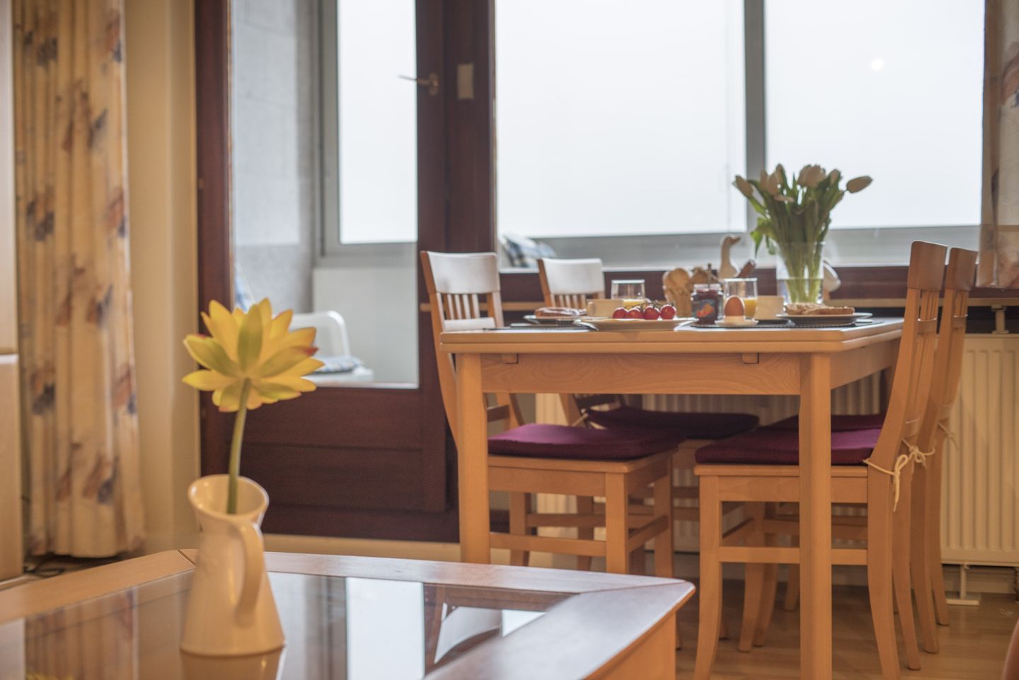 A dining area with a wooden table, chairs, a vase with flowers, and a window with curtains.