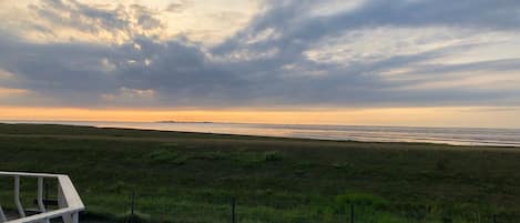 A beach view with a cloudy sky, a grassy area, and a paved walkway.