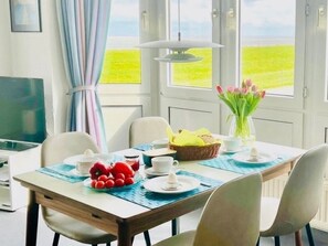 A dining area with a table set for two, a laptop, a basket of bread, and a vase of flowers.