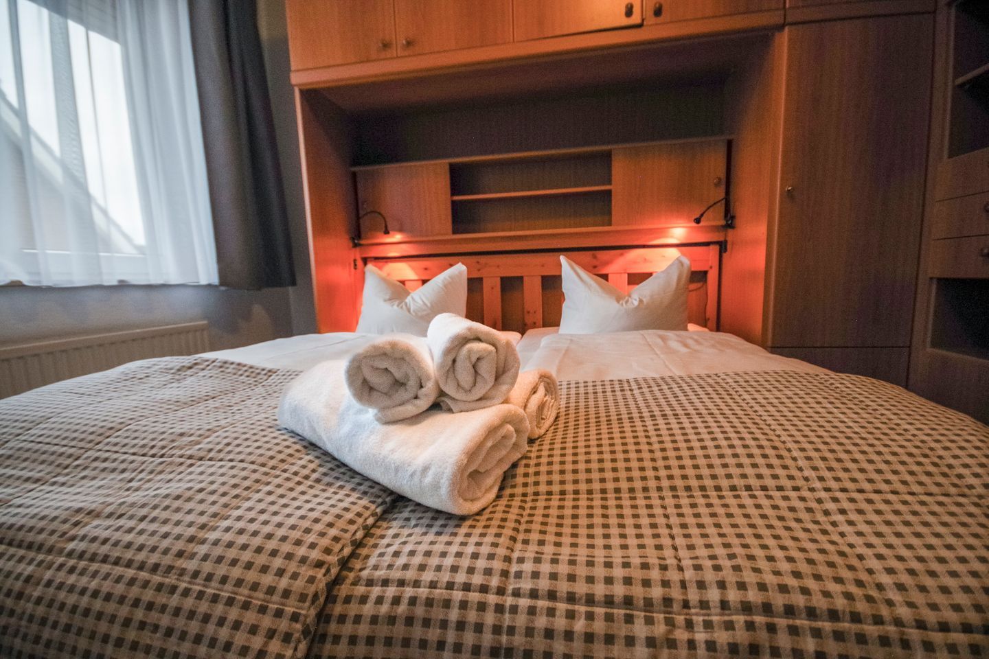 Ein ordentlich bezogenes Bett mit aufgerollten Handtüchern, einer karierten Tagesdecke und einem hölzernen Kopfteil.