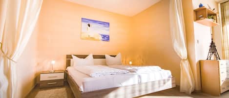 A bedroom with a bed, bedside tables, a nightstand, a painting, a dresser, and a window with curtains.