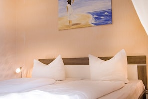 A neatly made bed with white linens and pillows, a wooden headboard, and a painting of a woman by the sea on the wall.