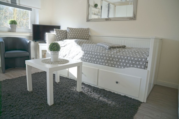 A bedroom with a bed, a small table with a plant, a chair, a TV, and a window with a view of trees.