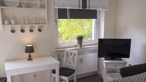 Une pièce avec un bureau blanc, une chaise, un téléviseur et une fenêtre donnant sur un paysage verdoyant.