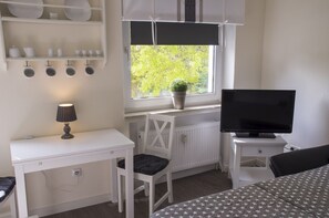Ein Zimmer mit weißem Schreibtisch, einem Sessel, einem Fernseher und einem Fenster mit Blick auf Grünflächen.