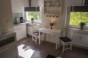 Una cocina compacta con muebles blancos, una mesa de comedor, sillas, un radiador, una ventana con vista a follaje, y una estantería con vajilla y tazas.