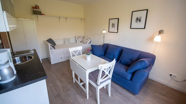 Un espacio habitable compacto con kitchenette, un sofá azul, una mesa de comedor blanca y un sofá pequeño blanco.