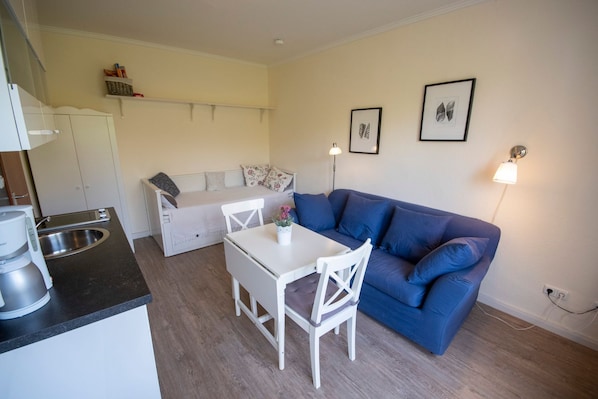 Un espacio habitable compacto con kitchenette, un sofá azul, una mesa de comedor blanca y un sofá pequeño blanco.