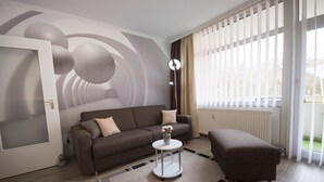 Un salon moderne avec un canapé, un pouf et une petite table sur laquelle est posé un vase. On y trouve une grande fenêtre avec des stores, un radiateur et une porte dotée d’un panneau en verre dépoli.