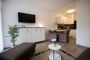 A compact living space with a kitchenette, a flat-screen TV mounted on the wall, a dining area with two chairs, and a small round table with a vase of flowers. - Fp104 - Strandnahe Ferienwohnung für 2 Personen, mit Schwimmbad-und Saunanutzung - Apartmenthaus (Cuxhaven)