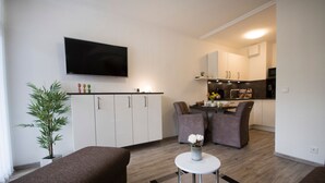 Un espace de vie compact comprenant un coin cuisine, un téléviseur à écran plat fixé au mur, un coin repas avec deux chaises et une petite table ronde agrémentée d’un vase de fleurs.