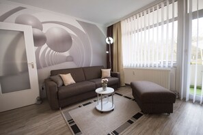 A modern living room with a sofa, ottoman, and a small table with a vase. There is a large window with blinds, a radiator, and a door with a frosted glass panel. - Fp104 - Strandnahe Ferienwohnung für 2 Personen, mit Schwimmbad-und Saunanutzung - Apartmenthaus (Cuxhaven)