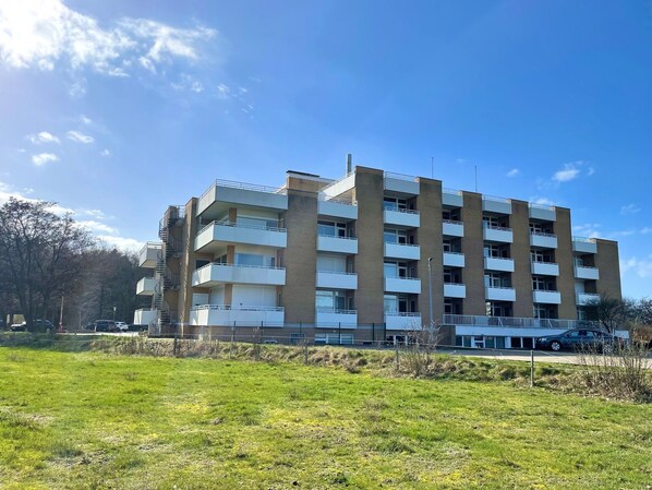 Un edificio residencial de varios pisos con balcones, un área verde al frente y un cielo azul despejado.