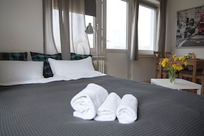 A hotel room with a bed, pillows, a desk, a chair, a vase with flowers, and a window with curtains.