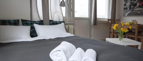 Hotelkamer met een bed, kussens, een bureau, een stoel, een vaas met bloemen en een raam met gordijnen.