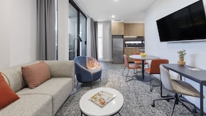 A modern hotel room with a sofa, armchair, dining table, chairs, and a flat-screen TV. - Punthill Essendon North (Essendon)