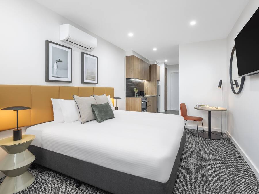 A modern hotel room with a large bed, a bedside table with a lamp, a small table with a chair, a TV mounted on the wall, and a kitchen area in the background.
