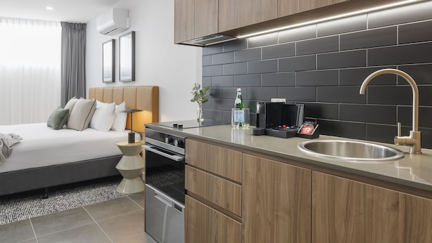 A modern hotel room with a bed, bedside table, and a kitchenette with a sink, stove, and microwave. - Punthill Essendon North (Essendon)