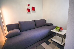 Uma sala de estar moderna com um sofá cinzento, uma pequena mesa lateral branca com uma tigela de frutas e três luzes vermelhas quadradas na parede.