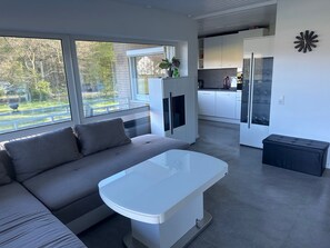 A modern living room with a sofa and a white coffee table, a kitchen with white cabinets, and a large window offering a view of the outdoors.