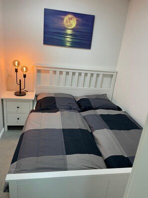 A bedroom with a white headboard, a bed with a grey and black checkered duvet, a nightstand with two lit lamps, and a painting of a moon over the sea on the wall.