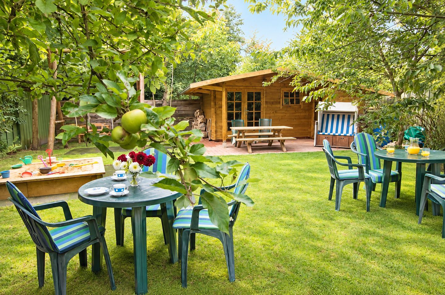 Een houten vakantiehuisje met een picknicktafel en stoelen, omgeven door een verzorgd gazon en bomen.