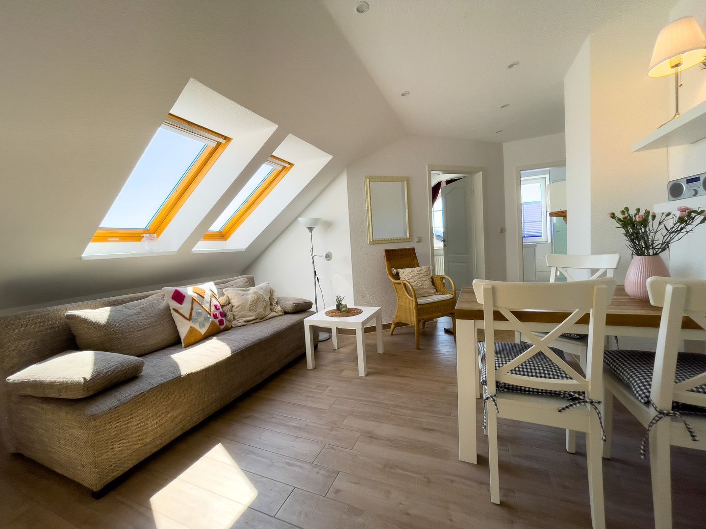 Ein modernes Wohnzimmer mit einer Couch, einem Esstisch und Stühlen. Im Deckenbereich befinden sich Oberlichter, eine Lampe und eine Vase mit Blumen.