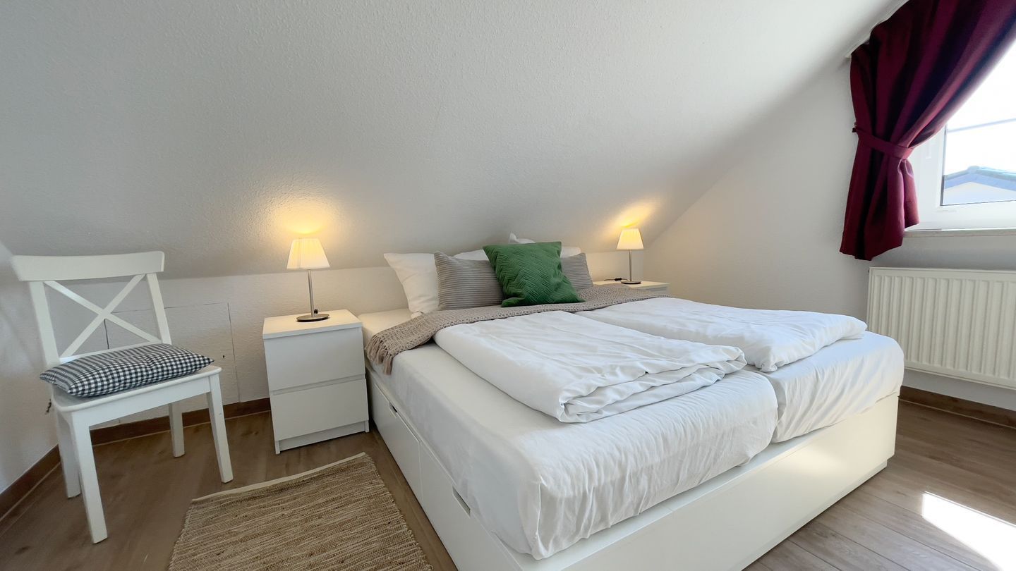 Ein Schlafzimmer mit einem Bett, zwei Nachttischen, einem Sessel und einem Fenster mit Vorhängen.