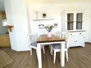 Un coin repas avec une table en bois, des chaises blanches, un buffet blanc aux portes vitrées et une petite étagère ornée de plantes et d’une radio.