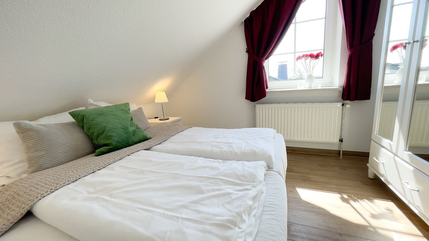 Ein Schlafzimmer mit zwei Betten, einem Heizkörper, einem Fenster mit Vorhängen und einem Nachttisch mit Lampe.