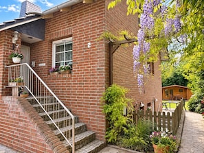 A brick house with a wooden porch, a staircase, and a garden with purple flowers.