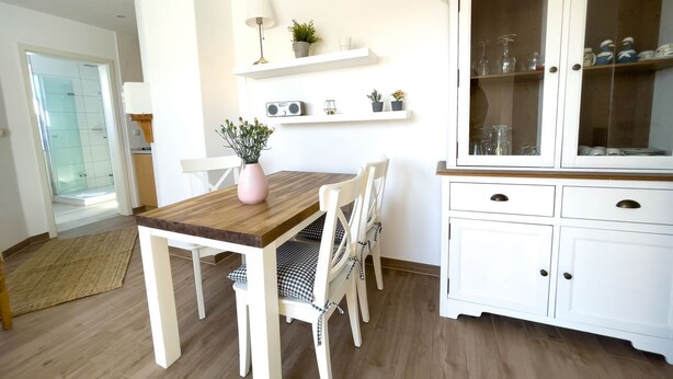 Een eethoek met een houten tafel en witte stoelen, een kast met glazen deuren, en een klein plankje met planten en een radio.