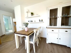 Un comedor con una mesa de madera y sillas blancas, un armario con puertas de cristal, y una pequeña estantería con plantas y un equipo de música.