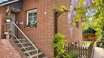 Ein Ziegelhaus mit einer Holzveranda, einer Treppe und einem Garten mit violetten Blumen.