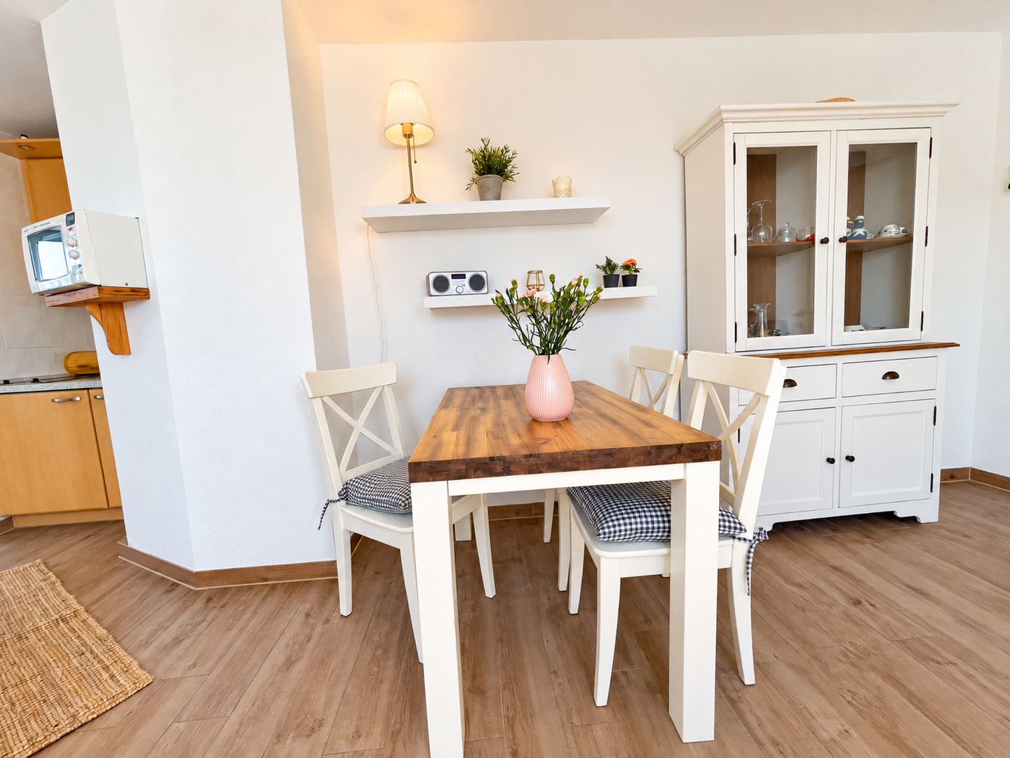 A dining area with a wooden table, white chairs, a white cabinet with glass doors, a radio on a shelf, and a small potted plant.
