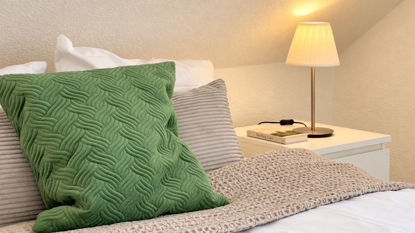 Una cama bien hecha con un almohadón verde con textura, una manta beige tejida y una lámpara de noche sobre una mesita blanca.