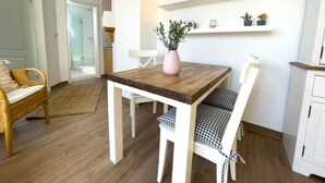 A dining area with a wooden table and white chairs, a wicker chair, a vase with flowers, and a kitchen in the background. - Kirsten 5 - Cozy vacation apartment close to the beach for up to 4 people - House (Cuxhaven)